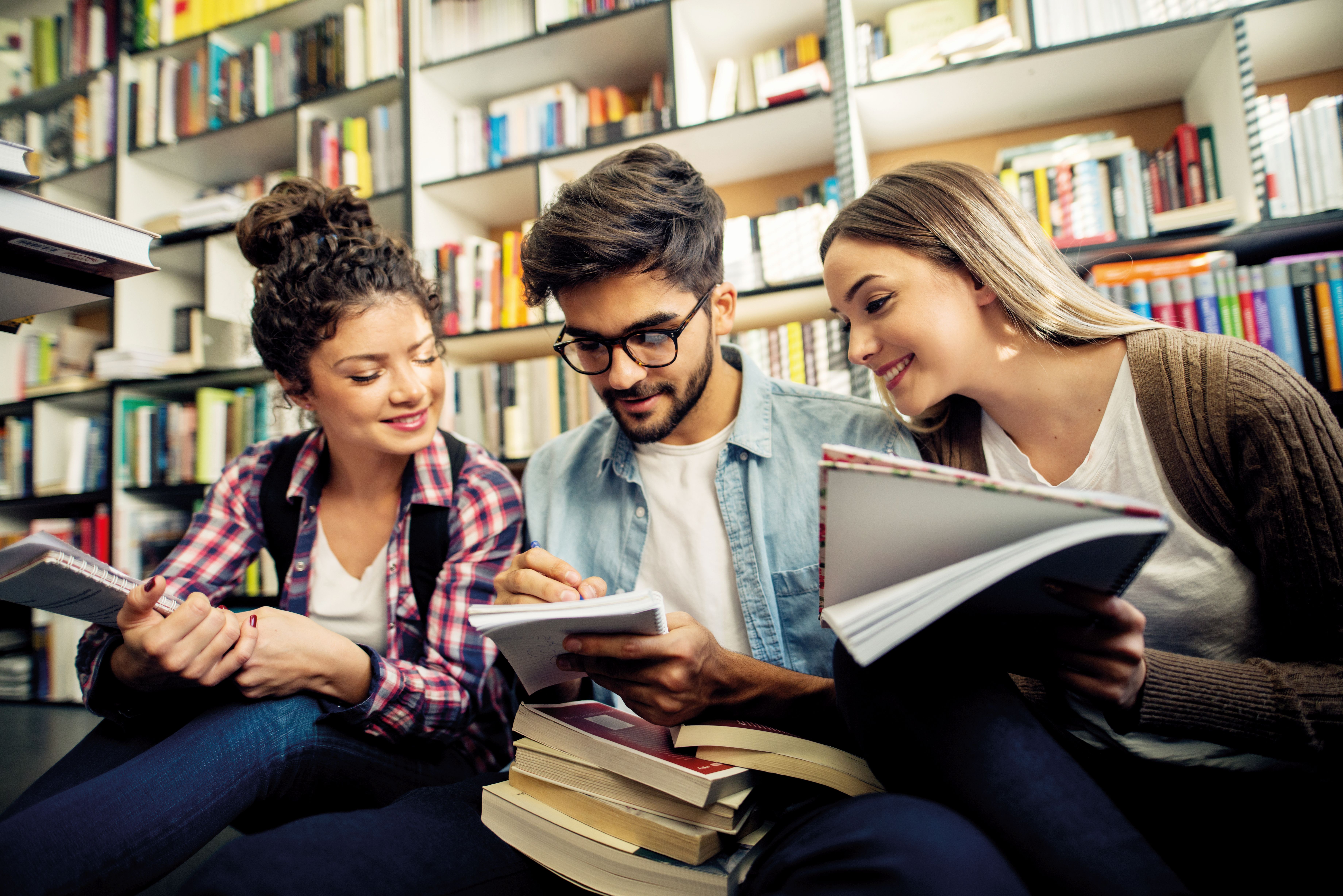 zwei junge Frauen und ein junger Mann sitzen vor einem Bücherregal und lesen